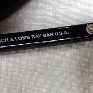 May include: Close-up of a black eyeglass frame with the text "BAUSCH & LOMB RAY-BAN U.S.A." in white. The frame is resting on a white textured surface. The hinge and screws are visible.