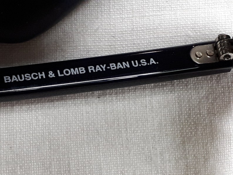May include: Close-up of a black eyeglass frame with the text "BAUSCH & LOMB RAY-BAN U.S.A." in white. The frame is resting on a white textured surface. The hinge and screws are visible.