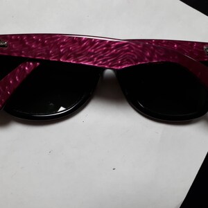 May include: A pair of black Ray-Ban sunglasses with a pink, textured frame. The sunglasses have a classic Wayfarer style.