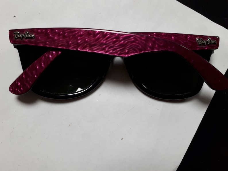 May include: A pair of black Ray-Ban sunglasses with a pink, textured frame. The sunglasses have a classic Wayfarer style.