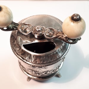 Pode incluir: Um cinzeiro antigo prateado com uma pega decorativa e dois botões cor de marfim. O cinzeiro tem formato redondo com padrões florais e de folhas. A pega é ornamentada com floreios decorativos.