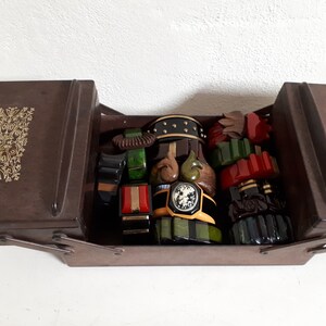 Könnte beinhalten: Eine braune Holzkiste mit einem Scharnierdeckel ist geöffnet und zeigt eine Sammlung von Vintage-Armbändern aus Kunststoff und Bakelit in verschiedenen Farben, darunter Grün, Rot, Schwarz und Braun. Die Armbänder sind gestapelt und in der Kiste angeordnet.