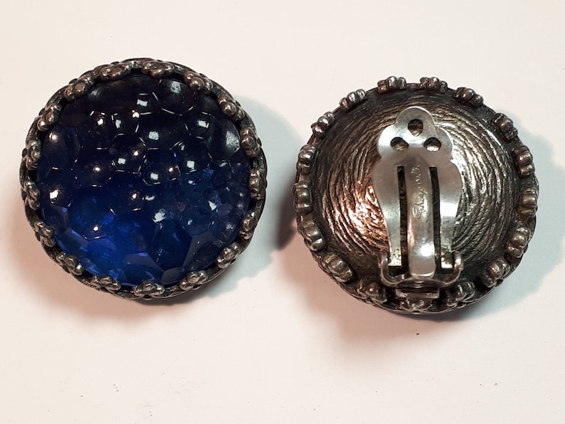 May include: Pair of vintage blue glass and silver tone clip-on earrings. The earrings feature a round blue glass cabochon set in a silver tone metal setting. The back of the earrings has a silver tone clip closure.