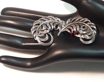 May include: A silver-toned brooch in the shape of two stylized feathers. The brooch has intricate detailing, with curved lines and a textured surface. The feathers are arranged side-by-side, creating a symmetrical design. The brooch is a vintage accessory.