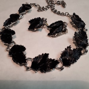Könnte beinhalten: Ein Vintage-Schmuckset mit einer Halskette und passenden Clip-Ohrringen. Die Halskette und die Ohrringe haben ein Blattdesign mit schwarzem Emaille und silberfarbenem Metall. Die Halskette ist mit einer Kette verstellbar.