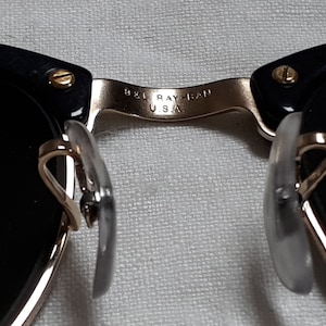May include: Close-up of black sunglasses with gold-colored accents. The bridge of the glasses has the text "B&L RAY-BAN U.S.A." The nose pads are clear and the lenses are black.