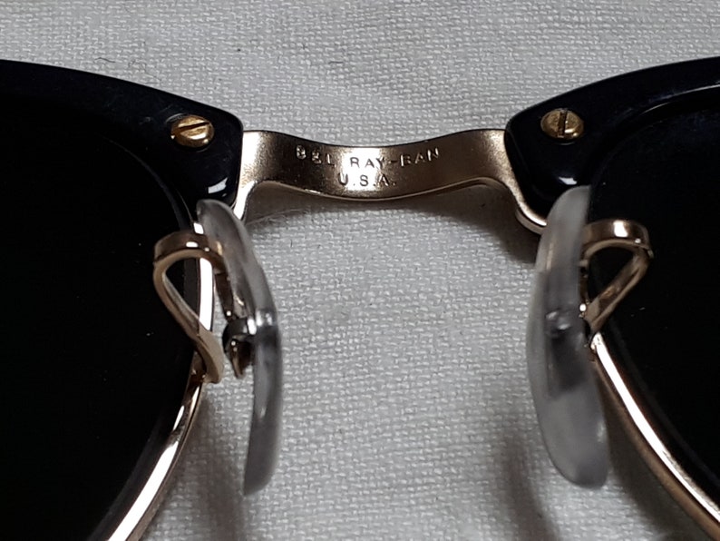 May include: Close-up of black sunglasses with gold-colored accents. The bridge of the glasses has the text "B&L RAY-BAN U.S.A." The nose pads are clear and the lenses are black.