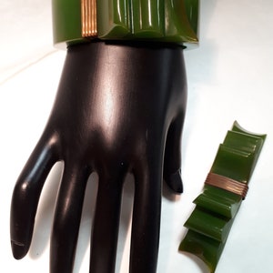 May include: A green and gold plastic bow bracelet and brooch set. The bracelet is on a mannequin hand and the brooch is on a white surface.