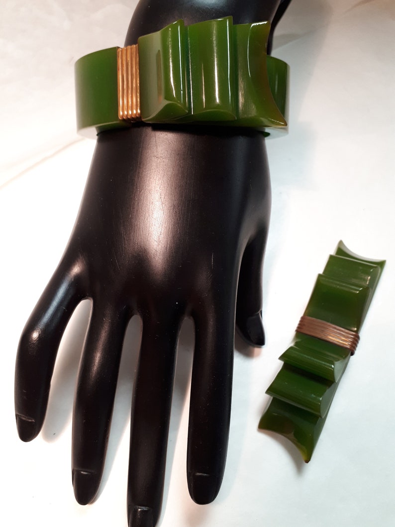 May include: A green and gold plastic bow bracelet and brooch set. The bracelet is on a mannequin hand and the brooch is on a white surface.