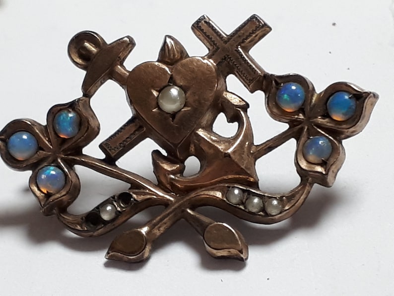 May include: A gold-toned brooch with a heart, anchor, and cross design. The brooch is decorated with opals and pearls.