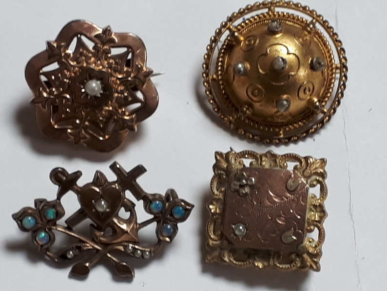 May include: Four antique gold brooches with pearl and opal accents. The brooches feature intricate designs, including a heart with crossed swords and anchors, a floral design, a round design with a raised center, and a square design with a raised center.