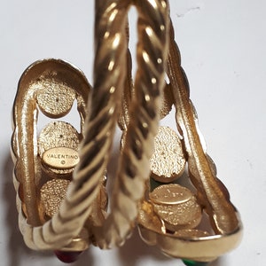 Puede incluir: Pulsera de brazalete de tono dorado con un dise&ntilde;o trenzado y piedras preciosas rojas y verdes. La pulsera est&aacute; marcada con "VALENTINO" en el interior.