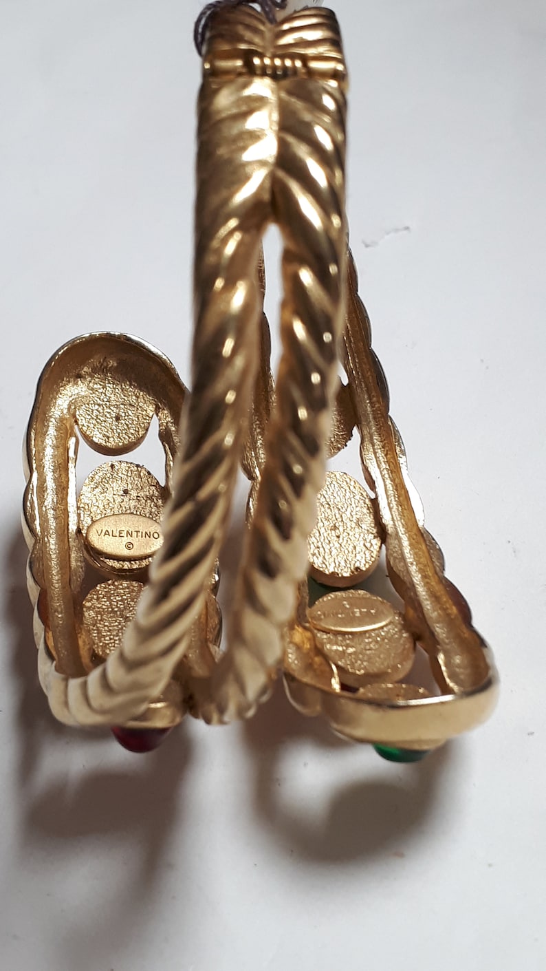 Puede incluir: Pulsera de brazalete de tono dorado con un dise&ntilde;o trenzado y piedras preciosas rojas y verdes. La pulsera est&aacute; marcada con "VALENTINO" en el interior.