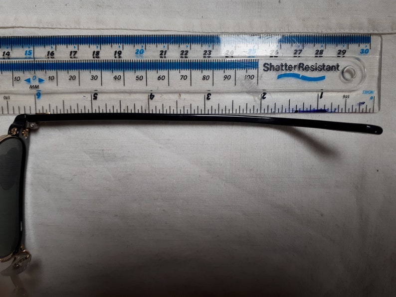 May include: Black sunglasses with a gold frame and dark lenses. The temple arm is measured with a ruler, showing a length of approximately 5 inches. The ruler is labeled "Shatter Resistant".
