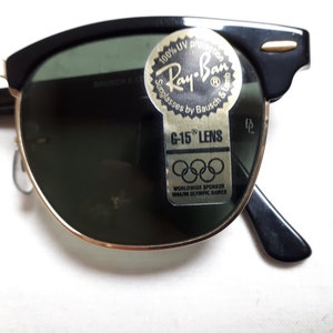 May include: Black and gold-rimmed Ray-Ban sunglasses with green lenses. The sunglasses have a classic design with a black upper frame and gold accents. A sticker on the lens reads "Ray-Ban Sunglasses by Bausch & Lomb" and "G-15 Lens."