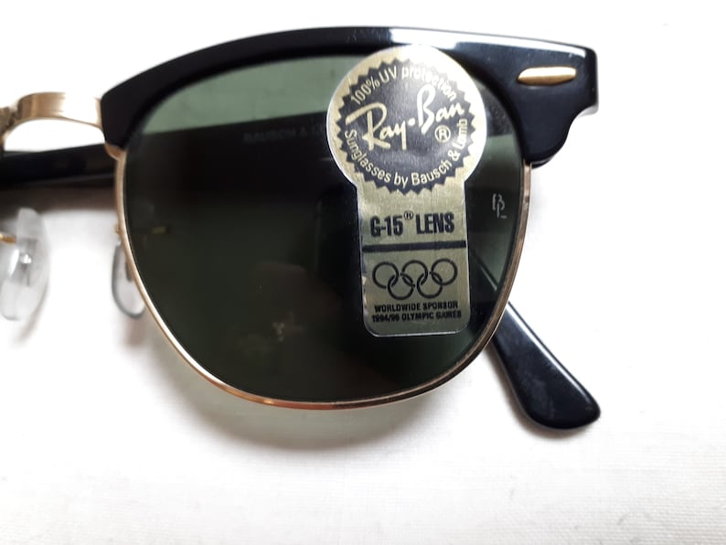 May include: Black and gold-rimmed Ray-Ban sunglasses with green lenses. The sunglasses have a classic design with a black upper frame and gold accents. A sticker on the lens reads "Ray-Ban Sunglasses by Bausch & Lomb" and "G-15 Lens."