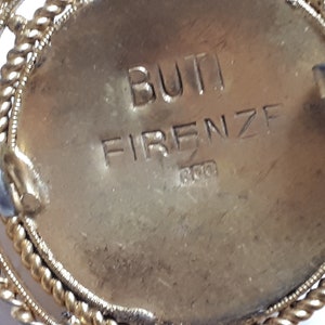 May include: Close-up of the back of a gold-toned brooch with a circular design. The brooch has a clasp and the inscription "BUTI FIRENZE 800" is engraved on the back.
