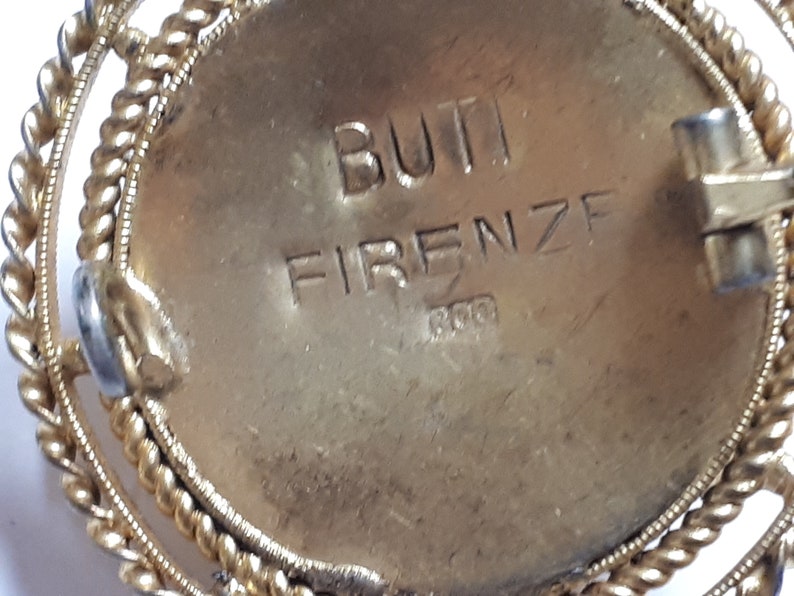May include: Close-up of the back of a gold-toned brooch with a circular design. The brooch has a clasp and the inscription "BUTI FIRENZE 800" is engraved on the back.