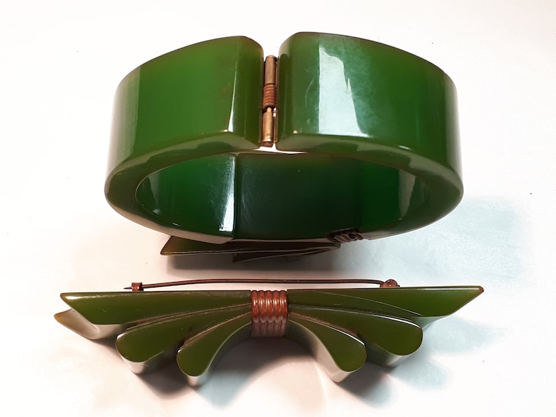 May include: A set of two vintage green Bakelite jewelry pieces. A wide green bangle bracelet with a gold-tone clasp and a matching green bow brooch with a brown metal center.