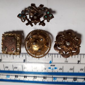 May include: Four antique gold-toned brooches with pearl accents. The brooches feature intricate designs, including a cross with floral accents, a round dome with a floral pattern, a square frame with an inscription, and a starburst design.