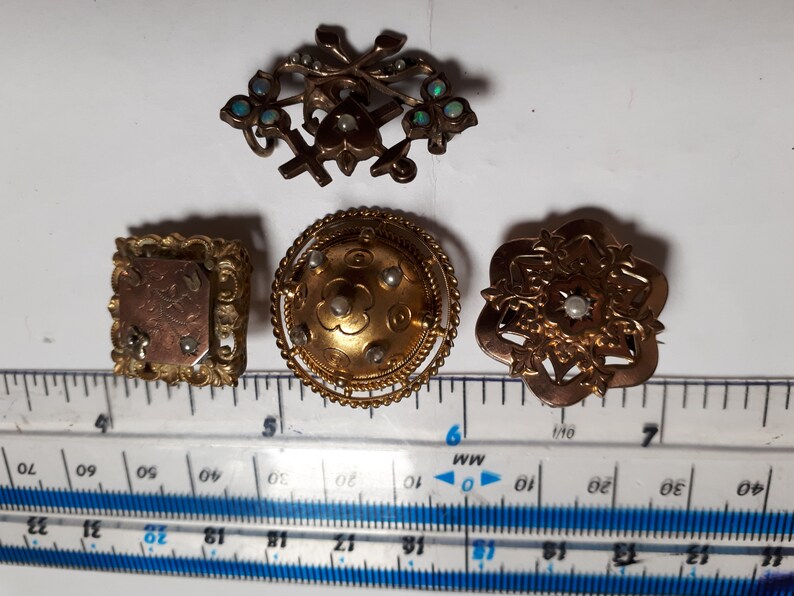 May include: Four antique gold-toned brooches with pearl accents. The brooches feature intricate designs, including a cross with floral accents, a round dome with a floral pattern, a square frame with an inscription, and a starburst design.