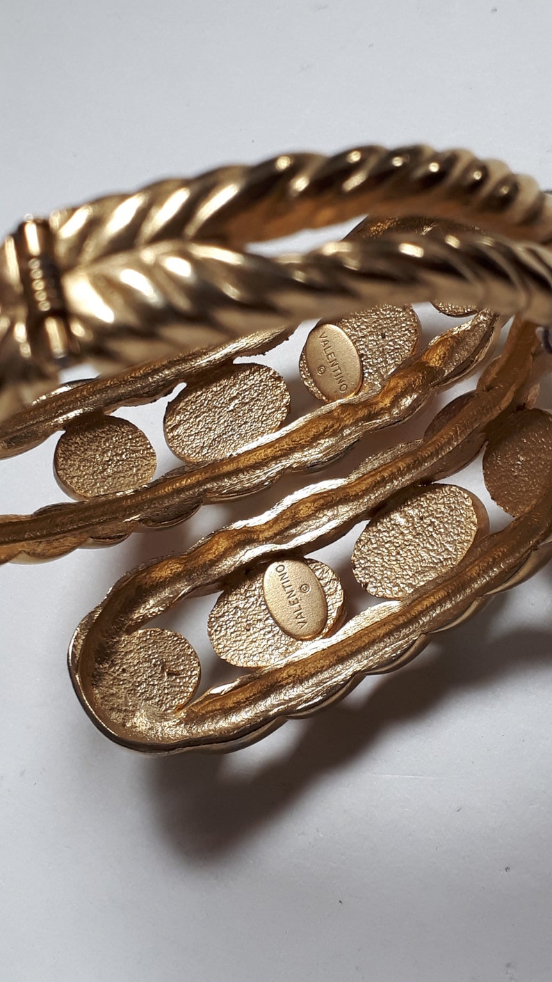 Puede incluir: Un brazalete de tono dorado con un dise&ntilde;o de cuerda trenzada y una serie de c&iacute;rculos redondos en relieve. El brazalete est&aacute; marcado con "VALENTINO JEWELRY" en el interior.
