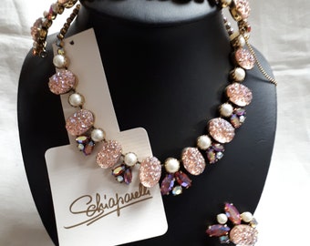 May include: A vintage jewelry set featuring a necklace, bracelet, and earrings. The set includes pink, iridescent, and pearl-like stones. The necklace and bracelet are displayed on a black mannequin head. The earrings are on the side. The brand name "Schiaparelli" is on a tag.