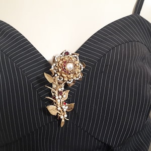 May include: A gold-toned brooch in the shape of a flower with red and white rhinestones. The brooch is pinned to a black and white striped fabric.
