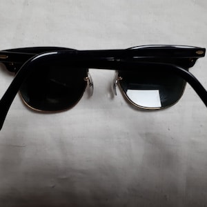 May include: Black sunglasses with a gold rim and dark lenses. The sunglasses have a classic design with a thick black frame and temples. The lenses are a dark green color.