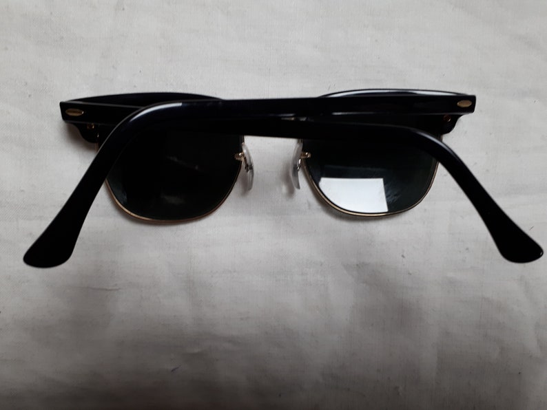May include: Black sunglasses with a gold rim and dark lenses. The sunglasses have a classic design with a thick black frame and temples. The lenses are a dark green color.