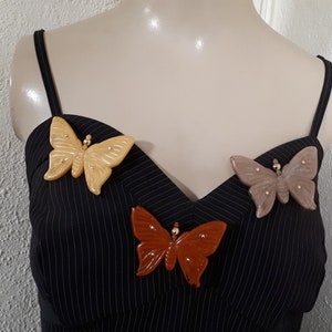 May include: Three butterfly brooches in different colors: beige, brown, and light brown. The brooches are made of a textured material and have gold accents.