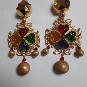 May include: A pair of gold tone dangle earrings with a four-leaf clover design. Each clover has four hearts in red, green, blue, and orange. The earrings have a gold ball at the bottom.