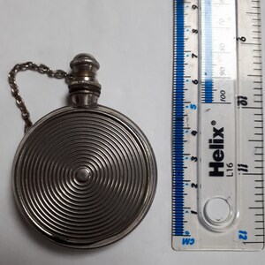 Può includere: Una bottiglia rotonda decorativa in argento con una catena e un piccolo tappo rotondo in argento. La bottiglia presenta un motivo a cerchi concentrici sul davanti. La bottiglia ha un diametro di circa 5 cm.