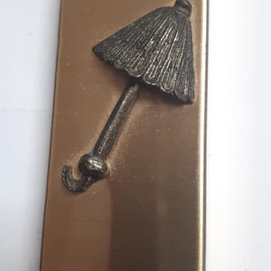 May include: A rectangular, bronze-colored metal object with a detailed, silver-colored umbrella design. The umbrella is embossed on the surface. A tan cord is attached at the top. The object has a brushed metal finish.