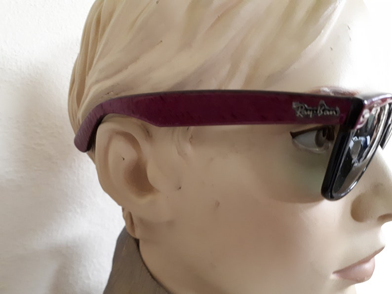 May include: A pair of dark purple Ray-Ban sunglasses with black lenses. The sunglasses are on a mannequin's head.