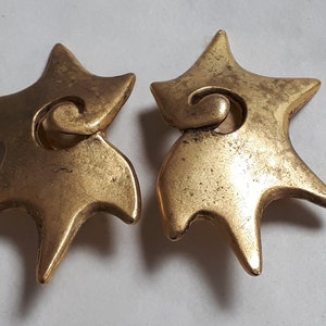 May include: A pair of gold-toned star-shaped earrings with a swirl design. The earrings are made of metal and have a slightly textured finish.
