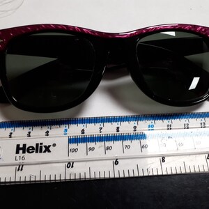 May include: Black sunglasses with a purple and black patterned frame. The sunglasses are measured against a ruler.