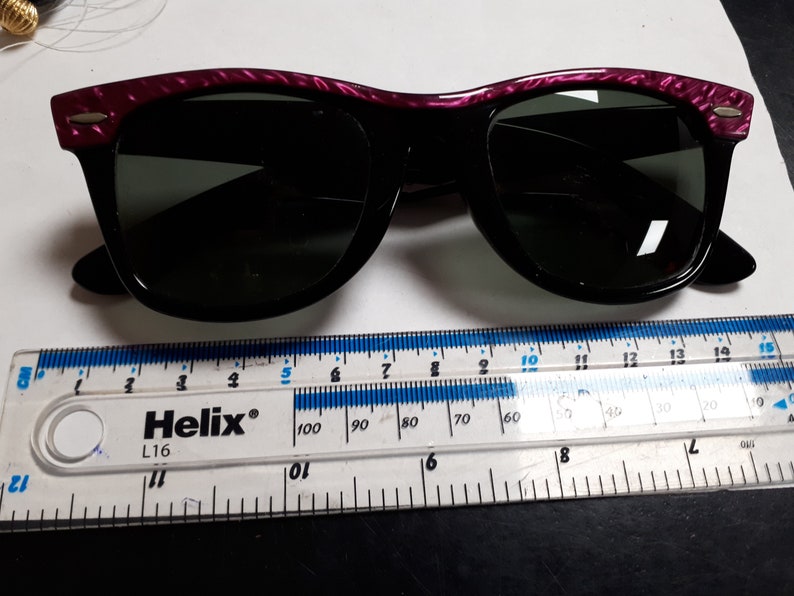 May include: Black sunglasses with a purple and black patterned frame. The sunglasses are measured against a ruler.
