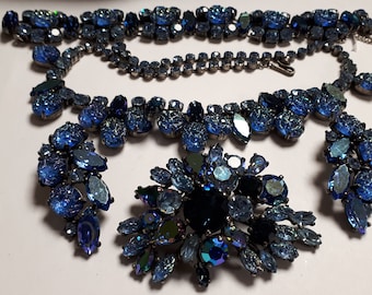 May include: A vintage blue rhinestone necklace and brooch set. The necklace features a large cluster of blue rhinestones in a teardrop shape. The brooch is a smaller version of the necklace, with a cluster of blue rhinestones in a flower shape.