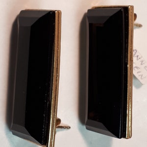 Puede incluir: Un par de pendientes rectangulares negros con ribetes dorados. Los pendientes están engastados con piedras negras.