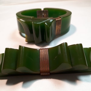 May include: A set of two green vintage bakelite jewelry pieces. The bracelet is a wide band with a bow design and a gold tone clasp. The brooch is a bow design with a gold tone center.