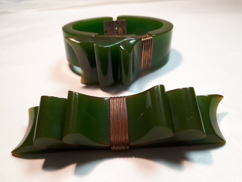 May include: A set of two green vintage bakelite jewelry pieces. The bracelet is a wide band with a bow design and a gold tone clasp. The brooch is a bow design with a gold tone center.