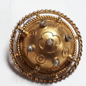 May include: A gold-toned brooch with a circular design. The center of the brooch features a raised, embossed flower with five petals. The flower is surrounded by a circle of smaller, embossed circles. The brooch is accented with six small, white stones.