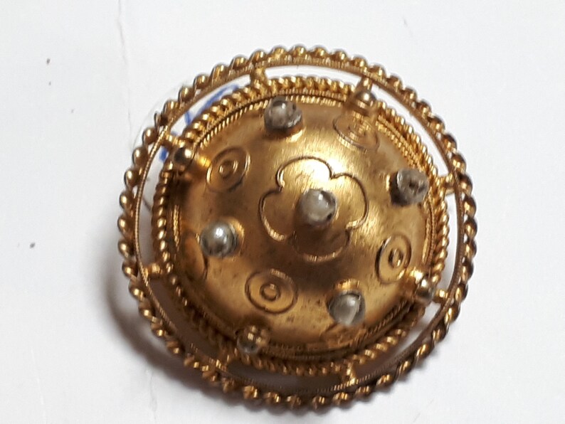 May include: A gold-toned brooch with a circular design. The center of the brooch features a raised, embossed flower with five petals. The flower is surrounded by a circle of smaller, embossed circles. The brooch is accented with six small, white stones.