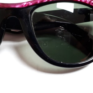 May include: A pair of black sunglasses with pink and silver frames. The lenses are dark green and have some scratches.