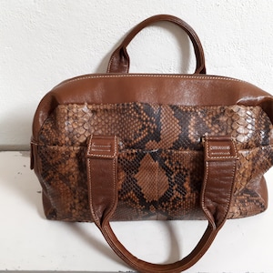 Könnte beinhalten: Eine braune Ledertasche mit Schlangenhautmuster. Die Tasche hat zwei obere Henkel und einen Reißverschluss.
