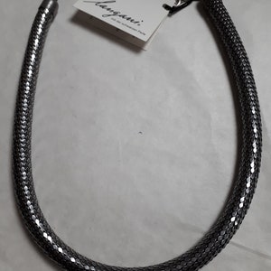 Puede incluir: Un collar de cadena de serpiente plateado con cierre de broche. La cadena está hecha de pequeñas escamas de metal entrelazadas. El collar está etiquetado con "Langani" y el texto "mit der schwarzen Perle".