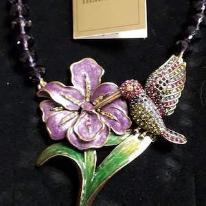 Könnte beinhalten: Eine lila Blume mit grünen Blättern und einem Kolibri, der darauf sitzt. Die Blume und der Kolibri sind aus Metall und haben eine goldene Oberfläche. Der Kolibri hat rote, grüne und blaue Emaille-Akzente. Die Halskette besteht aus schwarzen Perlen.