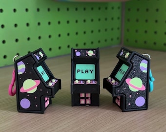 May include: Three black miniature arcade game keychains featuring a space theme with purple and lime green planets. The screen on the center unit displays the word PLAY in black text. Each unit includes a small metal ring and a colorful plastic clasp.