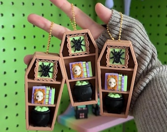May include: Three tan coffin-shaped ornaments with gold ball chains. Each features a black spider on a light green background, a skull on a book, a stack of books, and a black cauldron filled with light green liquid on the bottom shelf.